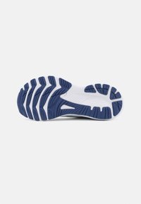 Semelle d'une chaussure de sport blanche et bleu marine avec des rainures profondes et des motifs d'adhérence sur un fond gris clair uni.