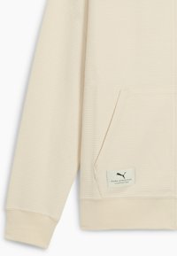 Cremefarvet, struktureret sweatshirtærme og lomme med et Puma Athletics-logo_patch nær lommens nederste kant.