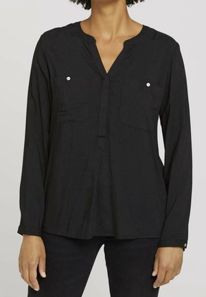 Femme portant un chemisier noir à manches longues avec deux poches poitrine et un col fendu, associé à un pantalon noir.
