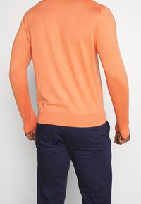 Orange långärmad tröja i mjukt stickat material med ribbade ärmslut och nederkant, i kombination med mörkblå byxor som har en figurnära design.