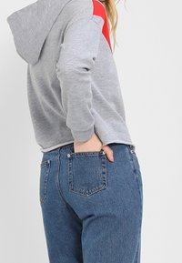 Szara bluza z kapturem z czerwonym akcentem na ramieniu, połączona z niebieskimi dżinsami. Bluza ma kaptur i luźny krój.