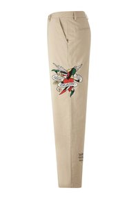 Beżowe bawełniane spodnie z haftem przedstawiającym ptaka, serce i wstęgę z napisem "PRAWDA ŚMIERCI". Zawierają subtelną czarną tekstową detal.