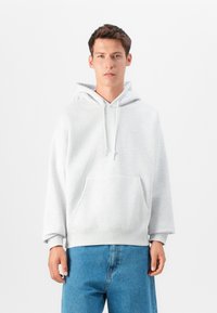 Sweatshirt cinza claro com capuz, feito de tecido macio, apresentando um bolso canguru e cordão, combinado com jeans denim descontraídos.