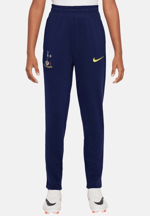 Marineblaue Sporthose mit elastischem Bund, gelber Nike-Swoosh auf dem rechten Oberschenkel, Sportteam-Logo auf dem linken Oberschenkel, getragen mit weißen Fußballschuhen.