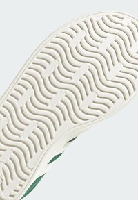 Suela de zapato de goma blanca con un patrón de olas y agarre texturizado. Material superior verde parcialmente visible a lo largo del borde, mostrando su diseño.