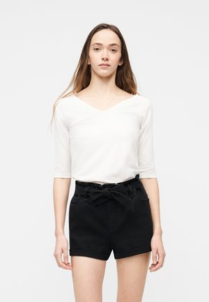 Ung kvinne med langt hår har på seg en hvit V-hals genser med albuelange ermer og høytlivs svarte shorts med knytt belte, stående mot en hvit bakgrunn.