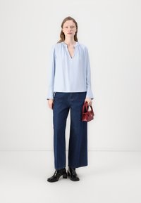 Blusa azzurro chiaro con scollatura a V e dettagli arricciati sul collo, abbinata a jeans a gamba larga blu scuro e stivali chunky neri. Tiene una piccola borsa rossa.