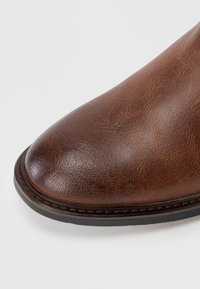 Bruine leren boot met een ronde neus, een gladde textuur, subtiel nerfpatroon en een zwarte rubberen zool. Steeksels bij de basis.