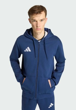 Jeune homme debout, les mains dans les poches, portant un sweat à capuche Adidas bleu marine et un pantalon assorti, sur un fond uni.