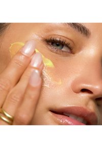 Nahaufnahme einer Hand, die ein gelbes Hautpflegeprodukt auf eine strahlende, glatte Gesichtsoberfläche aufträgt. Die Hand hat lange, natürliche Nägel.