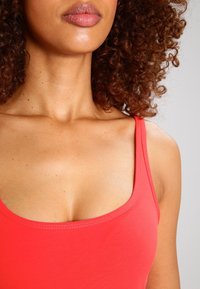 Canotta rossa con scollo a U, realizzata in tessuto liscio ed elastico, con sottili spalline e dettagli di cuciture discrete.