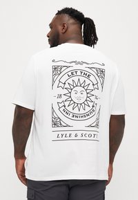 Valge lühikeste varrukatega t-särk, millel on julge must päikesegraafika koos tekstiga "LASKE PÄIKESEL SISE RONIDA" ja "LYLE & SCOTT" luksuslikus raamis.