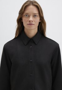 Donna con capelli castano chiaro che indossa una camicia nera con bottoni e colletto ampio, rivolta verso l'avanti su uno sfondo bianco semplice.