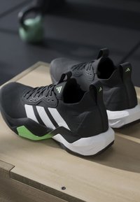 Paire de chaussures de sport noires avec des bandes blanches et des accents verts sur une surface en bois, matériel de gym flou en arrière-plan.