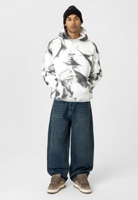Witte hoodie met zwarte abstracte patronen; gecombineerd met baggy blauwe jeans en beige hoge sneakers. Zwarte gebreide muts.