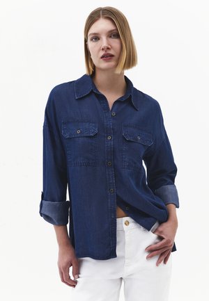 Kvinde med glat blond hår iført en mørkeblå denimskjorte med knapper og opsmøgede ærmer samt hvide bukser mod en hvid baggrund.