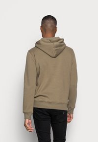 Sudadera de algodón marrón con una textura suave, bolsillo canguro y puños acanalados. Tiene capucha con cordón y un corte relajado. Pantalones negros visibles.