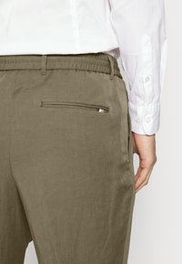 Olive-green pants with an elastic waistband, featuring a single back pocket with a striped accent. Smooth texture, tailored fit.
