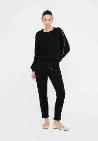 Zwarte gebreide pullover met versierde schouderdetails, gecombineerd met zwarte broek met trekkoord. Model draagt beige flats. Eenvoudige, ontspannen pasvorm.