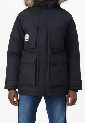 Chaqueta de plumas negra con capucha, que presenta un borde de piel sintética, múltiples bolsillos frontales y un parche redondo con el logo en el lado izquierdo del pecho.