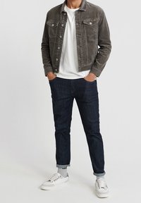 Giacca di velluto a coste grigia con due tasche sul petto, t-shirt bianca, jeans in denim scuro e sneakers bianche con dettagli grigi, in piedi su uno sfondo bianco.