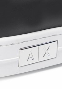 Black sneaker with a smooth texture, white rubber sole, and a silver metal logo plate displaying “A/X.”
