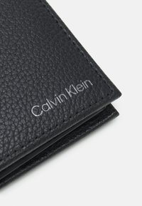 Fekete bőr pénztárca, texturált felülettel, ezüst "Calvin Klein" logóval, amely ki van nyomva az elején, és gondosan varrott szélekkel.