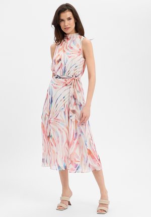 Frau trägt ärmelloses, pastellfarbenes Midi-Kleid mit abstraktem Muster, gebundenem Taillenschal und beigen Sandalen mit Absatz, steht vor weißem Hintergrund.