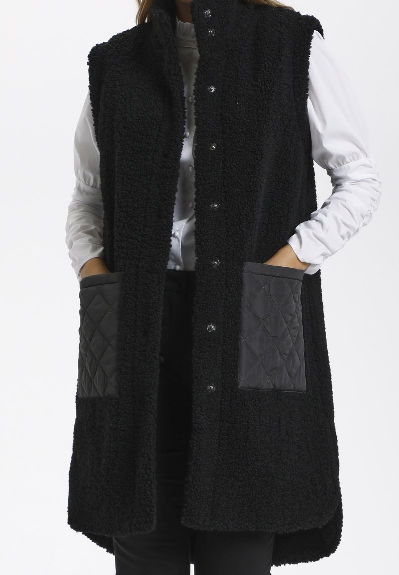 Femme portant un long gilet en polaire noir à boutons-pression et poches avant matelassées, par-dessus un chemisier blanc et un pantalon noir.
