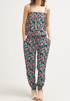 Woman wearing a black sleeveless floral jumpsuit with red and blue flowers, hands in pockets, paired with beige open-toe heels.