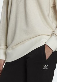 Person som bär en beige sweatshirt med muddar och svarta Adidas-byxor med en hand i fickan som visar Adidas-logotypen på låret.