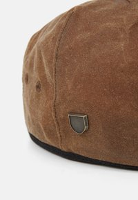 Casquette en toile cirée marron avec une forme arrondie, un large bord sombre et un petit badge en métal sur le côté. Surface texturée avec des coutures visibles.