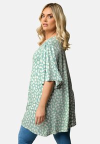 Ljusgrön tunika med vitt lövmönster, korta ärmar och lös passform, kombinerad med blå jeans. Mjuk tyg med draperad design.