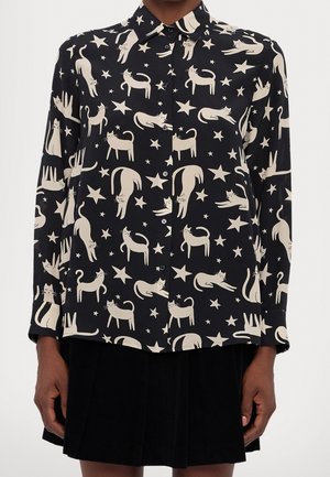 Sort skjorte med knapper med mønster af cremefarvede katte i forskellige poser og stjerner, båret med et sort skørt.