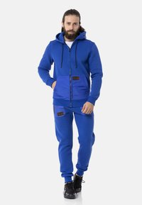 Blaue Kapuzenjacke und passende Jogginghose aus weichem Stoff. Mit Vordertaschen mit Lederakzenten und verstellbarer Kapuze mit Kordelzügen.