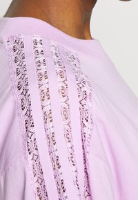 Ljusrosa t-shirt med spetsdetaljer längs axeln och horisontella ränder, tillverkad av mjukt tyg.