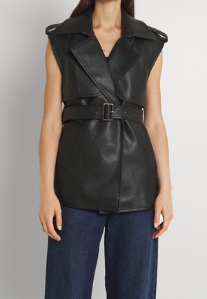 Ärmellose schwarze Lederweste mit breiten Revers und Gürtel, getragen über dunkelblauer Jeans, gezeigt vom Hals bis zu den Hüften.