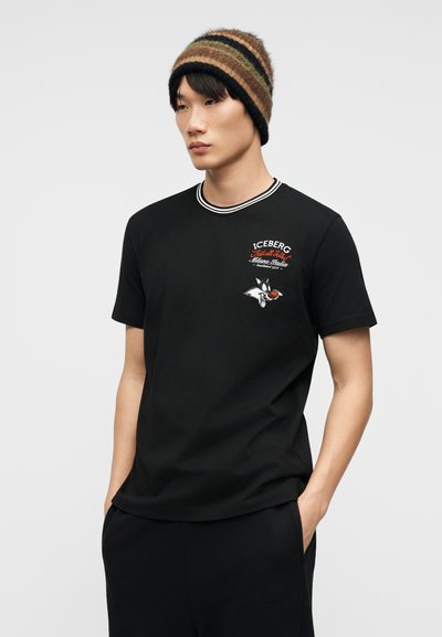 Νεαρός άνδρας που φοράει ριγέ καφέ σκουφάκι και μαύρο T-shirt με το λογότυπο "Iceberg" και καρτούν χαρακτήρα στο στήθος, με τα χέρια στις τσέπες, κοιτώντας προς τα αριστερά.