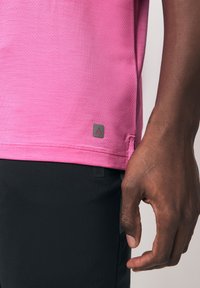 Mano dalla pelle scura appoggiata di lato, che indossa una maglietta rosa a maniche corte dalla texture e pantaloni neri con piccoli tag a forma di triangolo su entrambi gli indumenti.