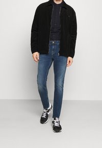 Zwarte corduroy jas met kraag, marineblauwe button-up shirt, slim-fit blauwe jeans en zwarte sneakers. De jas heeft een ritsluiting en een gestructureerde stof.