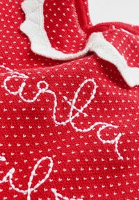 Roter Strickstoff mit weißen Punkten, verziert mit weißer Stickerei, die Text zeigt, sowie einem gewellten Saum am Ausschnitt.