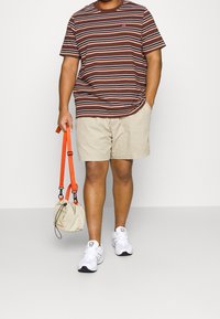 Camiseta de algodón a rayas en marrón y blanco, pantalones cortos beige, zapatillas blancas y un pequeño bolso beige con correas naranjas.