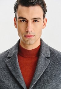 Szary wełniany płaszcz z klapami z wycięciami, noszony na czerwonym golfsze z prążkowanego materiału. Zbliżenie podkreśla fakturę tkaniny i precyzyjne detale krawiectwa.