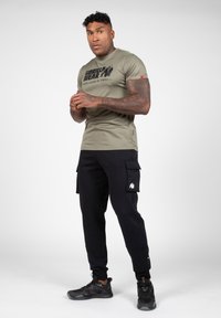 Olijfgroene aansluitende T-shirt met zwart "GORILLA WEAR" logo, gecombineerd met zwarte cargobroek met zijzakken en een grote witte graphic.