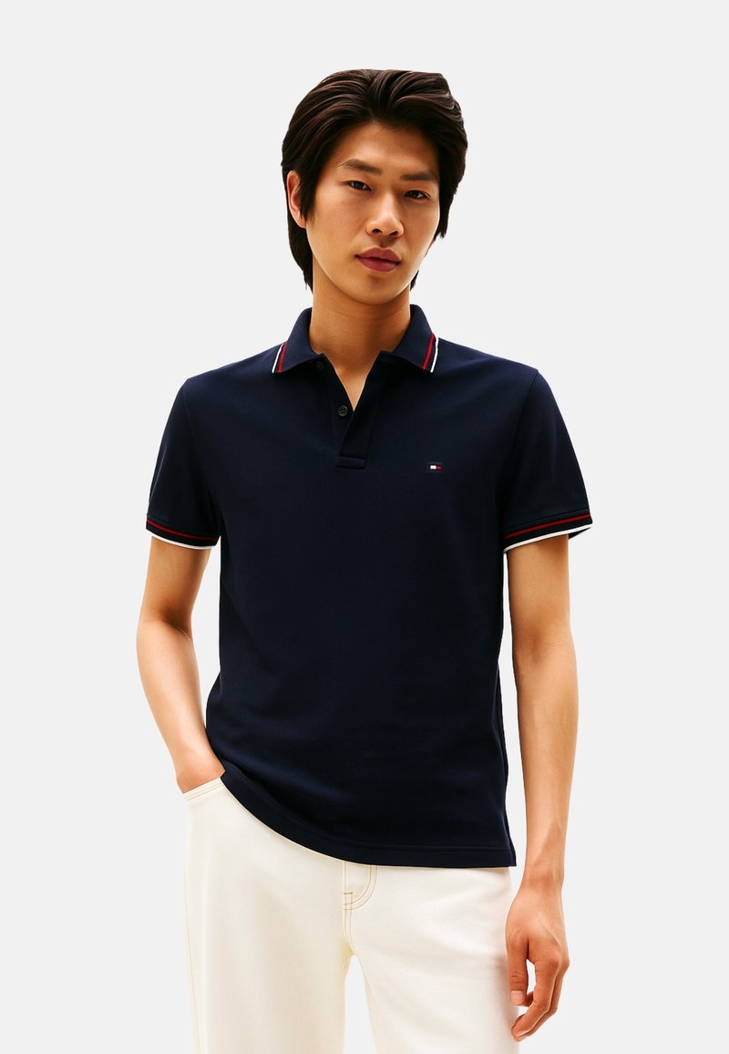 Camisa polo azul marino con cuello que presenta detalles en rojo y blanco. Mangas cortas y diseño ajustado. Pequeño logo en el pecho.