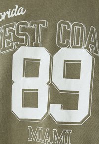 Tejido verde oliva con texto en blanco. Presenta "FLORIDA" en la parte superior, "COSTA OESTE" y un gran número "89", con "MIAMI" debajo. Textura suave.