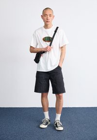 Hvit t-skjorte med grønn og brun spiraldesign, sorte denimshorts, svart crossover-veske, svart og hvite sneakers med hvite sokker.