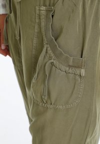 Primer plano de un bolsillo de pantalones verde oliva con cordón y ribete de punto acanalado en la cadera y mano de una persona.