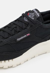 Zapatilla negra con parte superior de lona, con detalles cosidos, marca Reebok y suela de goma texturizada en color crema. Diseño elegante con cordones.