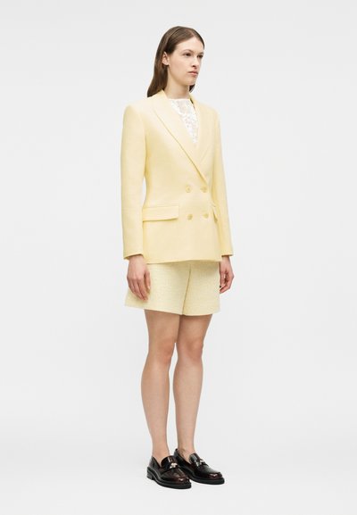 Femme debout portant un blazer croisé jaune pâle, un short texturé crème, un haut en dentelle blanche et des mocassins noirs brillants sur un fond blanc.